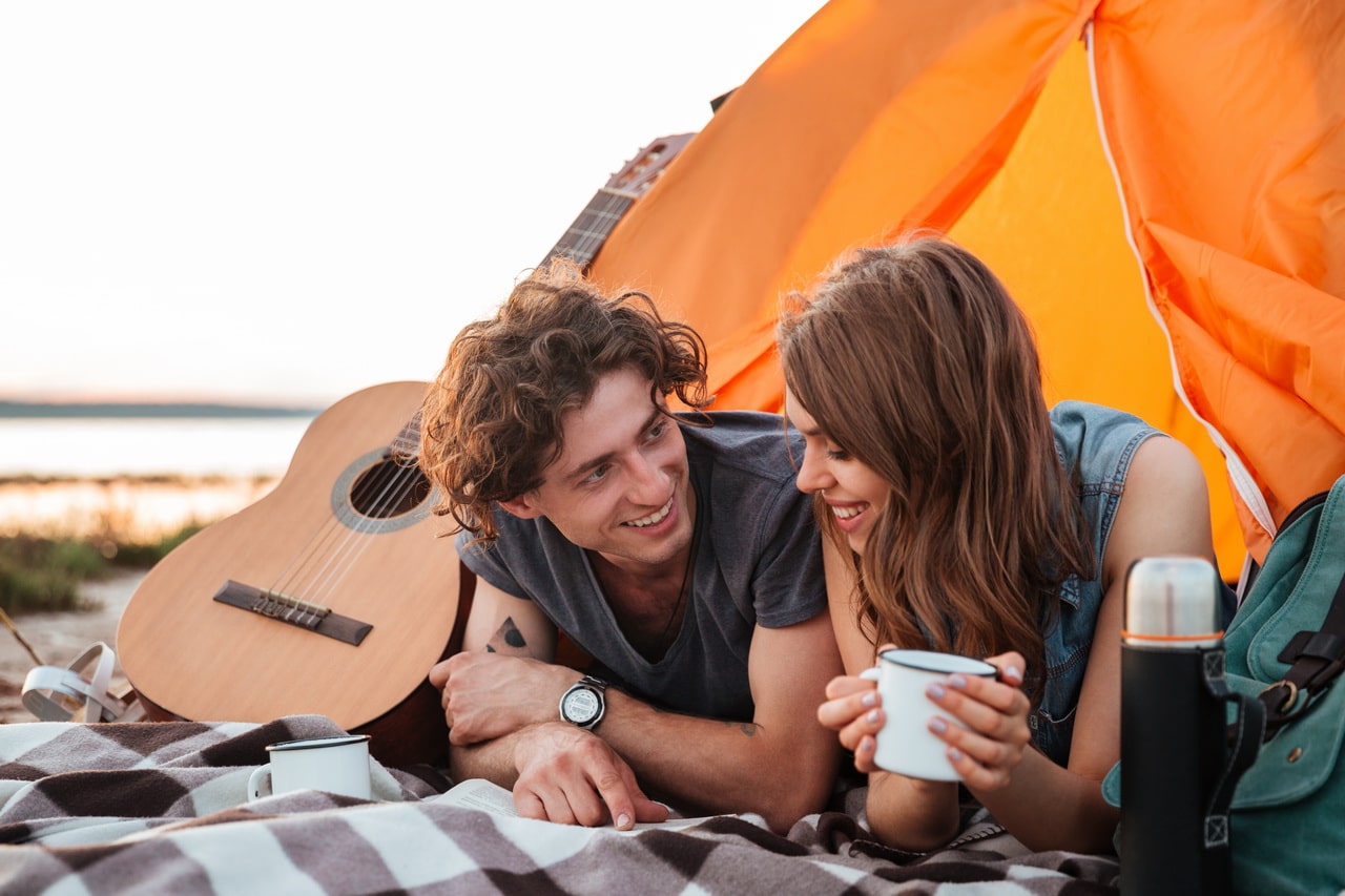 Happy Young Couple Lying And Drinking Tea In Touristic Tent