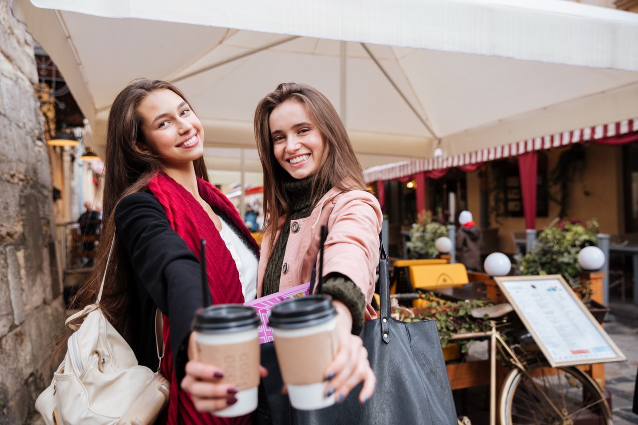 Two Cheerful Young Women Holding Cups Of Coffee To Go